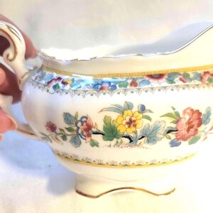 May include: A cream-coloured porcelain creamer featuring a scalloped rim and a gold-trimmed handle. The body is decorated with a colourful floral pattern in shades of yellow, pink, and blue, accented by a yellow and gold geometric border near the top.