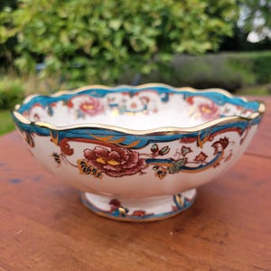 May include: A white ceramic bowl with a scalloped edge, adorned with a floral pattern in red, teal, and gold. The bowl rests on a small pedestal base, displaying a classic decorative design.