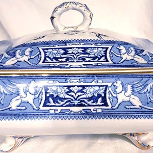 May include: A rectangular ceramic serving dish with a lid, featuring a white base with intricate blue patterns of winged lions and floral motifs. The piece includes gold-toned trim on the edges, handles, and feet, with a central loop handle on top.
