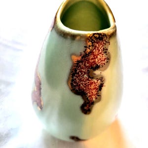 May include: A teardrop-shaped ceramic vase with a smooth, pale mint green glaze. The surface features irregular, textured patches of deep reddish-brown and metallic copper-toned oxidation, with a gold-toned rim around the opening.