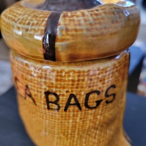May include: A ceramic tea bag holder in the shape of a burlap sack. The holder is tan with the word "BAGS" in black lettering. The lid is brown and has a dark brown stripe down the side.