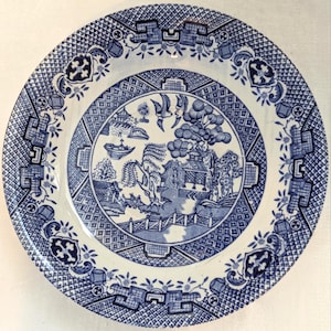 May include: A round ceramic plate featuring a blue and white willow pattern. The central design depicts a landscape with a bridge, a pagoda, trees, and two birds in flight. The border is decorated with a geometric lattice and floral motifs.