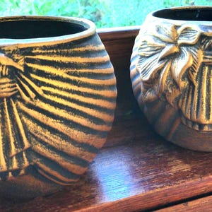 May include: Two decorative, round, gold-coloured vases with a black interior. Each vase features a detailed relief design of a bow and radiating lines. The vases are displayed on a wooden surface.