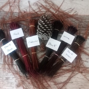 May include: Bundles of natural pine needles dyed in various colors, including red, natural, black, wine, dark blue, and dark green. The bundles are arranged on a wooden surface.