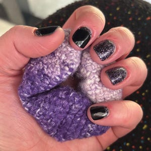 Könnte beinhalten: Eine Hand hält eine gehäkelte Haargummi. Der Haargummi hat einen Farbverlauf aus lila und weißem Garn. Die Nägel sind schwarz lackiert mit silbernen Glitzerakzenten.