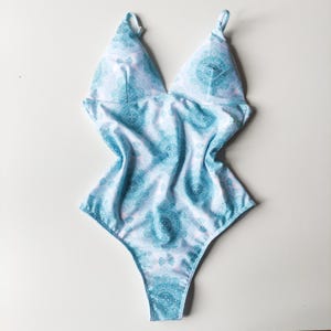 Può includere: Un costume da bagno blu chiaro con un motivo a mandala bianco e blu. Il costume da bagno ha una scollatura profonda e spalline sottili.