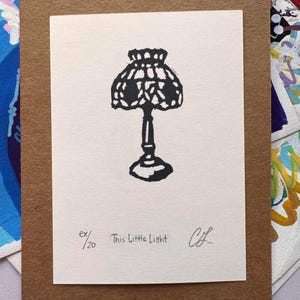May include: A black ink block print on a cream-colored rectangular card featuring a lamp illustration. Below the lamp, the text reads "ex/20", "This Little Light", and a handwritten signature. The card is mounted on a brown paper backing.