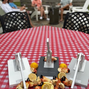 May include: Three crab crackers on a red and white checkered tablecloth with crab claws and lemons scattered around them. The middle cracker has a black base and the text "Garden Cottage" engraved on it.