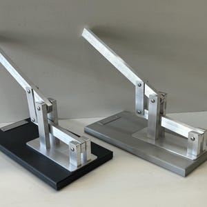 May include: Two silver metal lever-action devices on black and gray rectangular bases. The levers are raised and the devices appear to be in a closed position.