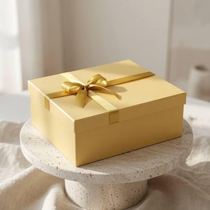May include: A rectangular gift box in a soft, pale yellow hue, featuring a matching satin ribbon tied in a bow on the lid. The box is positioned on a circular, speckled, off-white stone pedestal with a smooth, flat top and a cylindrical base.