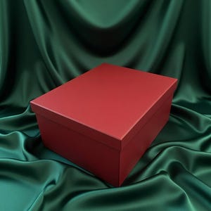 Könnte beinhalten: Eine rote rechteckige Box mit Deckel, vor einem Hintergrund aus drapiertem smaragdgrünem Stoff. Die Box hat eine glatte, matte Oberfläche. Der Stoff hat eine seidige, fließende Textur.