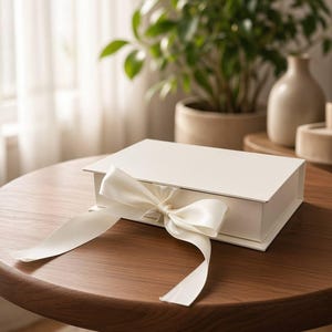 May include: A rectangular white gift box with a smooth matte finish, featuring a wide, off-white satin ribbon tied into a decorative bow at the front center. The box has a hinged lid design and sits flat, showcasing a clean, minimalist aesthetic.