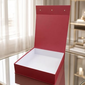 May include: A square, deep red gift box with a hinged lid that stands upright. The exterior of the box is a solid matte red, while the interior base and the inside of the lid are finished in a clean, contrasting white. Three small circular magnets are visible.