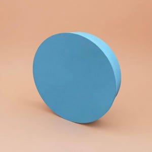 Puede incluir: Un disco circular de color azul claro sólido con un acabado mate y suave. El objeto está colocado en ángulo, mostrando su cara frontal plana y redonda junto a un borde cilíndrico grueso. El color es un azul cielo vibrante y uniforme.