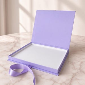 Pode incluir: Uma caixa de presente retangular na cor lavanda com tampa articulada aberta. O interior da caixa é branco e uma fita de cetim lavanda correspondente está presa à borda frontal, estendendo-se para fora para permitir fechar a caixa.