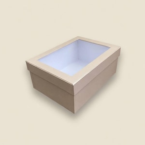 Op de afbeelding: Een rechthoekige beige doos met een doorzichtig venster op het deksel. De doos heeft een witte binnenkant en is ontworpen voor verpakking of opslag. De doos is gemaakt van karton of een vergelijkbaar materiaal.
