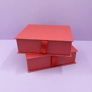 Può includere: Due scatole regalo quadrate di colore rosso opaco impilate l'una sull'altra. Ogni scatola presenta un coperchio piatto e una linguetta in nastro rosso abbinato centrata sul bordo anteriore, progettata per facilitare l'apertura.