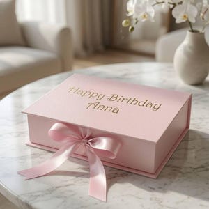 May include: A rectangular light pink gift box with a hinged lid, featuring the gold embossed text Happy Birthday Anna on the top surface. The box is secured with a matching light pink satin ribbon tied in a bow at the front center.