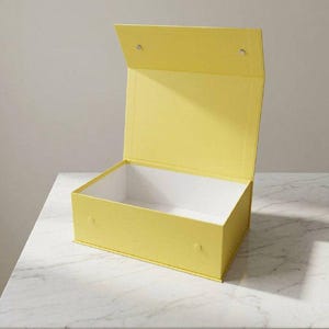 Op de afbeelding: Een rechthoekige gele geschenkdoos met een open scharnierend deksel. De buitenkant van de doos is effen felgeel van kleur, terwijl de binnenkant wit is. Twee kleine ronde magnetische sluitingen zijn zichtbaar aan de voorzijde en het deksel.