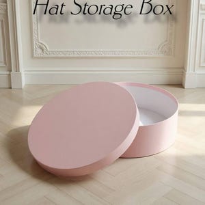 Puede incluir: Una caja rosa para guardar sombreros con la tapa parcialmente abierta, revelando el interior blanco. La caja redonda está sobre un suelo de madera clara. El texto "Hat Storage Box" está encima.
