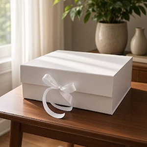 Op de afbeelding: Een vierkante witte geschenkdoos met een gladde, matte afwerking, voorzien van een scharnierend deksel en een decoratief wit satijnen lint dat aan de voorzijde in het midden tot een strik is gestrikt. De doos is gemaakt van stevig materiaal.