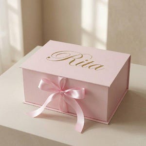 Könnte beinhalten: Eine rechteckige, hellrosa Geschenkbox mit Klappdeckel, auf dem der Name Rita in eleganter goldener Schreibschrift aufgedruckt ist. Die Vorderseite der Box ist mit einem passenden hellrosa Satinband verziert, das mittig zu einer Schleife gebunden ist.