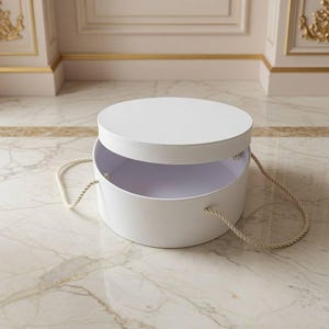 May include: A round white box with a matching lid and two beige twisted rope handles attached to the sides. The box is open, revealing a hollow interior, and the lid is positioned slightly above the base, showing a clean and simple design.