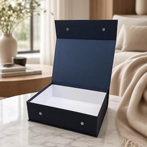 Può includere: Una scatola rettangolare blu navy con coperchio incernierato in posizione verticale. L'interno della scatola è bianco. L'esterno presenta due piccole chiusure magnetiche circolari color argento sulla patta anteriore e due sul coperchio.