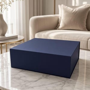 Può includere: Una scatola regalo quadrata blu navy opaco con coperchio incernierato. La scatola presenta un esterno pulito e in tinta unita con bordi netti e una finitura liscia, presentata come un contenitore chiuso per la conservazione o l'esposizione.