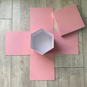 Könnte beinhalten: Eine zerlegte rosa Geschenkbox mit sechseckigem Innenraum und Deckel. Die Box ist geöffnet und zeigt das weiße Innere des sechseckigen Fachs. Die Box befindet sich auf einer Holzoberfläche.