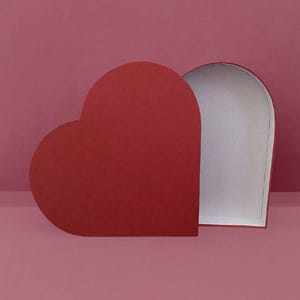Puede incluir: Una caja roja en forma de corazón, parcialmente abierta, que revela un interior blanco. La caja está colocada sobre un fondo rosa, creando un esquema de color monocromático. La caja es probablemente para regalos o almacenamiento.