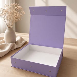 Pode incluir: Uma caixa de presente quadrada de cor violeta claro com uma tampa articulada que permanece na vertical. O exterior da caixa é de uma cor lavanda sólida, enquanto o interior é branco. Três pequenos fechos magnéticos prateados são visíveis.