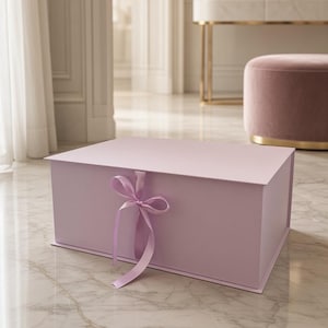 Op de afbeelding: Een rechthoekige, lichtlavendelkleurige geschenkdoos met een scharnierend deksel en een bijpassend satijnen lint dat aan de voorzijde in het midden tot een strik is gestrikt. De doos heeft een gladde, matte afwerking en is van stevig materiaal.