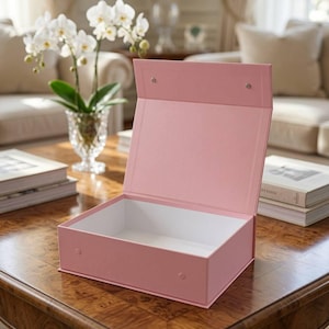 May include: A rectangular, light pink gift box with a hinged lid that is open. The exterior of the box is a solid matte pink, while the interior is white. Two small, circular magnetic closures are visible on the front edge and the lid.