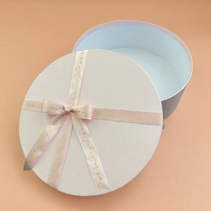 May include: A round, white gift box with a matching lid. The lid is decorated with a light pink ribbon tied in a bow, featuring gold script text and small heart icons. The interior of the box is visible, showing a light blue lining inside the container.