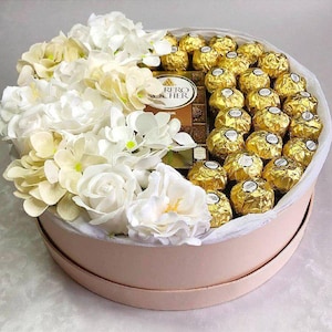 Pode incluir: Uma caixa de presente redonda rosa claro cheia de flores brancas e creme, chocolates embrulhados em ouro e uma caixa de chocolates Ferrero Rocher. A caixa é forrada com papel branco.