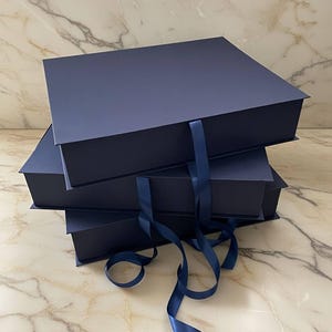 Könnte beinhalten: Drei marineblaue Geschenkboxen, die übereinander gestapelt und mit passendem Satinband gebunden sind. Die quadratischen Boxen haben einen Magnetverschluss. Das dunkelblaue Band ist zu sehen.