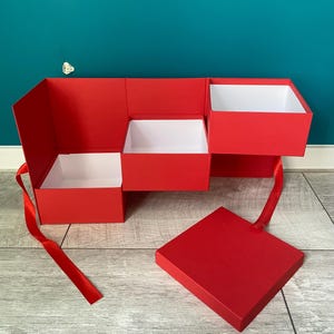 Könnte beinhalten: Eine rote Geschenkbox mit mehreren Fächern und einem roten Band. Die Box ist geöffnet und die Fächer sind sichtbar. Die Box steht auf einer Holzoberfläche.