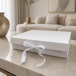 Puede incluir: Una caja de regalo cuadrada de color blanco con tapa abatible y una cinta de satén blanca atada en un lazo en el centro de la parte frontal. La caja tiene un acabado liso y mate con un diseño limpio, ofreciendo una presentación sencilla.