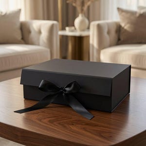 May include: A rectangular black gift box with a hinged lid and a black satin ribbon tied in a bow at the front center. The box has a smooth, matte finish and is constructed from rigid material, presenting a clean and structured appearance.