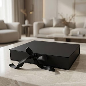 May include: A square, matte black gift box with a hinged lid, featuring a black satin ribbon tied in a bow at the front centre. The box has a clean, structured design with sharp edges and a smooth, solid black finish on all visible exterior surfaces.