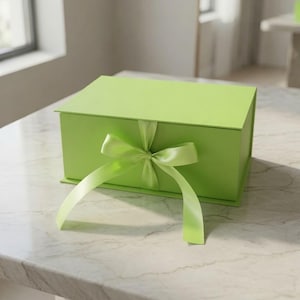 Pode incluir: Uma caixa de presente retangular verde-limão com tampa articulada e uma fita de cetim verde-limão a condizer, atada num laço na parte frontal central. A caixa tem um acabamento mate liso e está posicionada horizontalmente.