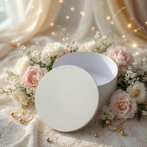 Può includere: Una scatola regalo rotonda bianca con il coperchio abbinato posizionato davanti, circondata da un gruppo di rose rosa pallido e bianche, piccoli fiori bianchi e diversi piccoli ciondoli sferici dorati su un tessuto di pizzo bianco.