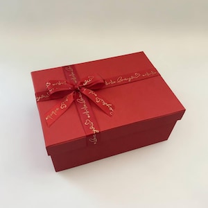 May include: A red gift box with a matching ribbon tied in a bow. The ribbon has gold text and small heart designs. The box is rectangular and sits on a white surface.