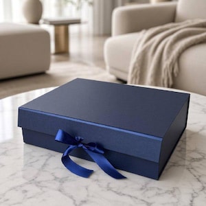 Può includere: Una scatola regalo rettangolare blu navy con una finitura opaca liscia, dotata di un nastro di raso blu navy abbinato legato a fiocco sul pannello frontale. La scatola è chiusa e presenta un design pulito e strutturato con bordi netti.