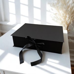 May include: A rectangular black gift box with a matte finish, featuring a hinged lid and a black satin ribbon tied in a bow at the front center. The box is constructed from rigid material and maintains a clean, structured, and minimalist appearance.
