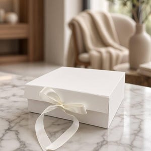 May include: A square white gift box with a smooth matte finish, featuring a hinged lid and a decorative white satin ribbon tied in a bow at the front center. The box is constructed from rigid material and maintains a clean, minimalist aesthetic.
