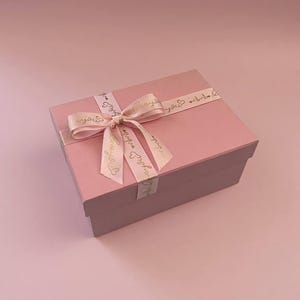 Pode incluir: Uma caixa de presente rosa com uma tampa a condizer e uma fita atada. A fita é num tom de rosa mais claro com texto dourado e um design de coração. A caixa está sobre uma superfície rosa.