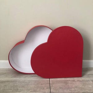 May include: A heart-shaped gift box with a vibrant red exterior and a clean white interior. The lid is detached and positioned to the right, revealing the hollow white cavity of the base. The box features smooth, solid-colored surfaces.