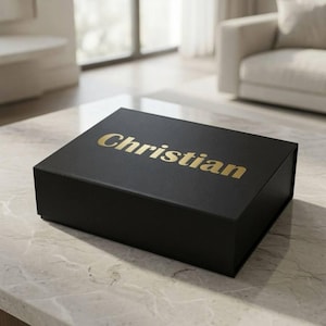 Puede incluir: Una caja de regalo rectangular de color negro con acabado mate que presenta la palabra Christian impresa en letras doradas en la tapa. La caja está cerrada y tiene un diseño estructurado con bordes definidos, ofreciendo un aspecto uniforme.
